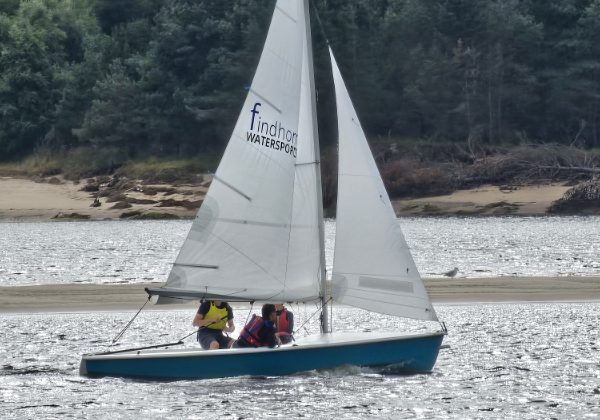 Sailboat with crew on shimmering water near forested shore